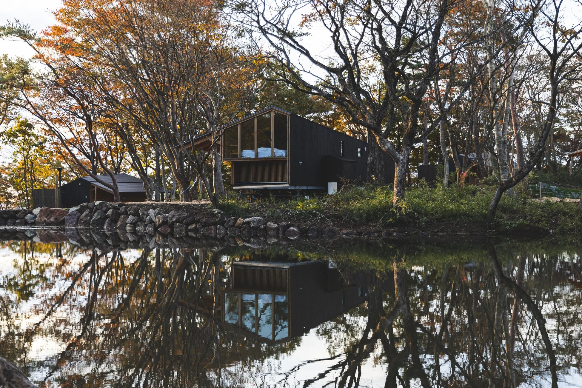 秋の紅葉に囲まれた湖畔に立つモダンな黒い家が、水面に美しく反射している風景。静かで落ち着いた雰囲気が漂っています。