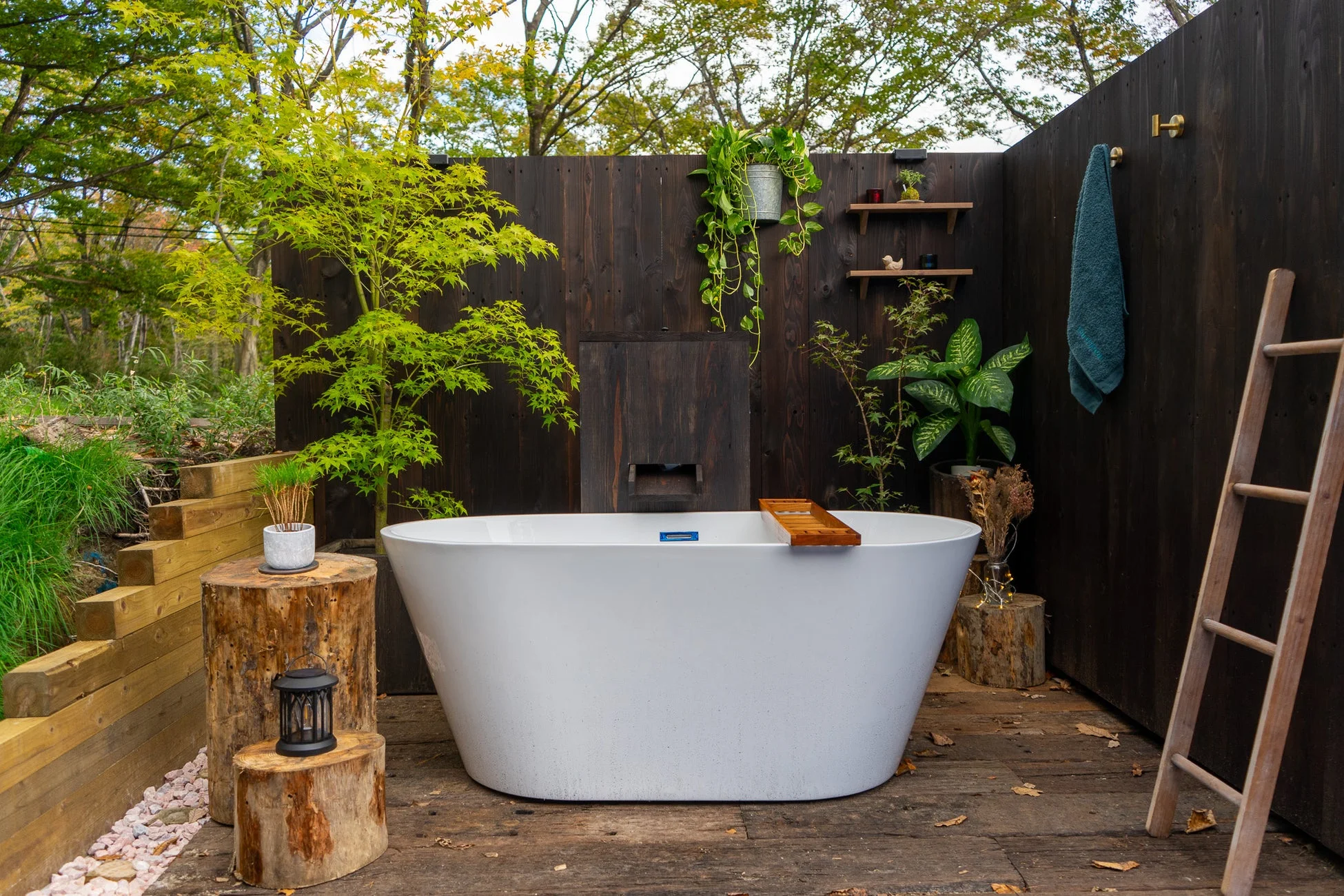 緑豊かな自然の中に設置された白い独立型浴槽が特徴の屋外バスルームです。木製の壁や床、切り株のサイドテーブルがナチュラルな雰囲気を醸し出し、リラックスできる空間を提供しています。