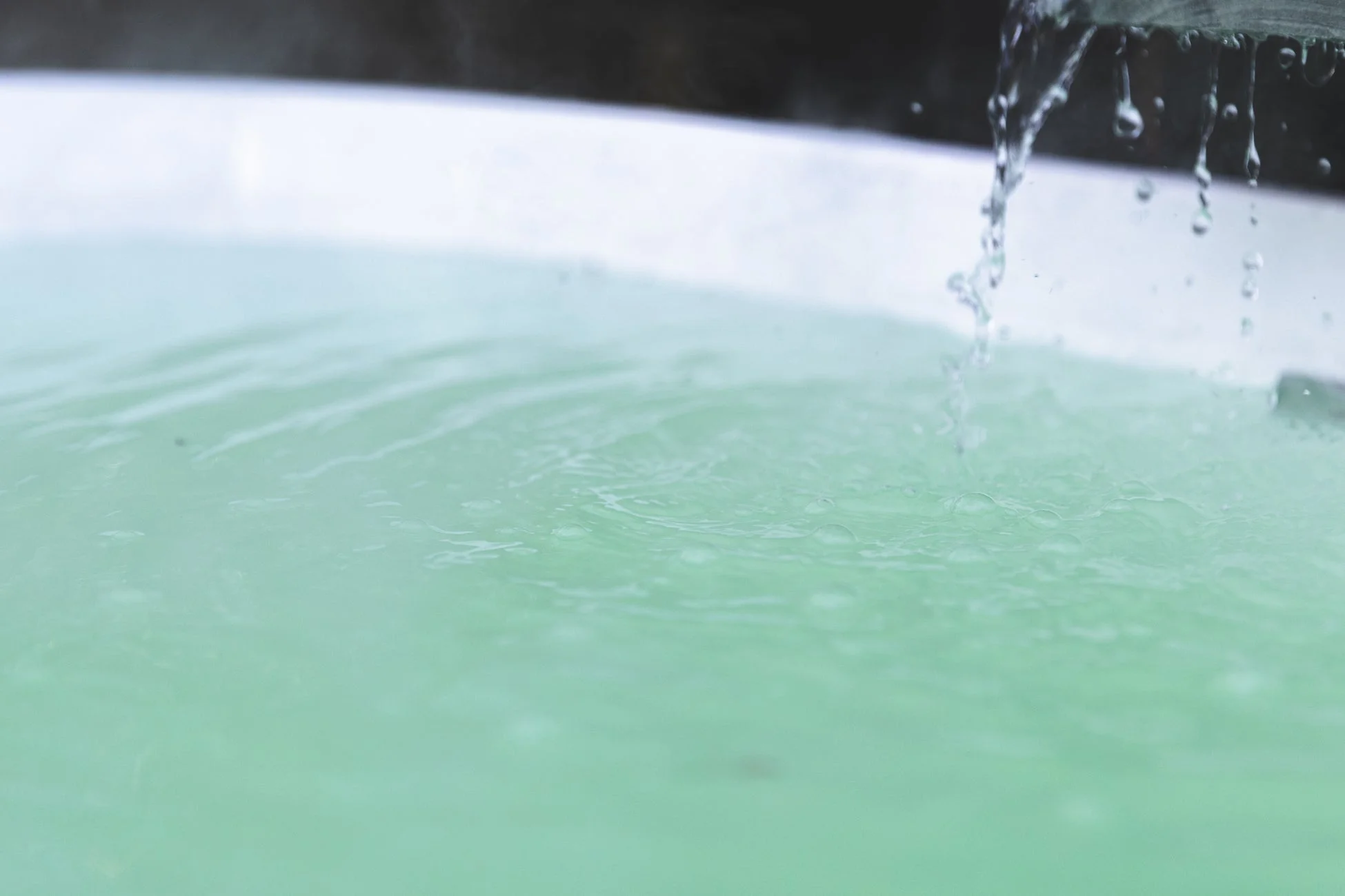 エメラルドグリーンの湯が張られた浴槽に、勢いよくお湯が注ぎ込まれている様子を捉えたクローズアップ。湯気も立ち上り、温かさと癒しを感じさせます。水面に広がる波紋と水滴が印象的です。
