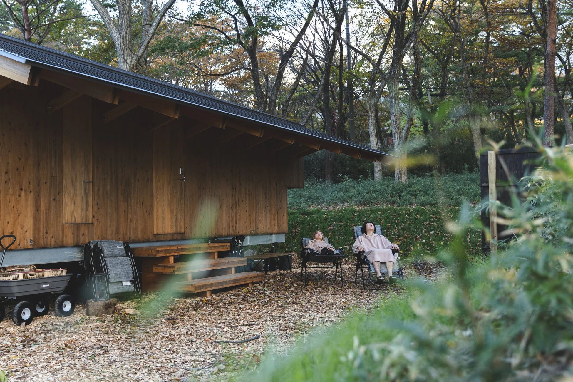 木造の建物の前で、二人がリクライニングチェアに座り、秋の自然の中でリラックスしている様子。落ち葉が敷き詰められた静かな空間で、ゆったりとした時間を過ごしている。